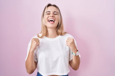 Pembe arka planda duran genç sarışın kadın başarı için heyecanlı kolları havada ve gözleri kapalı zaferi kutlarken gülümsüyor. Kazanan konsept. 