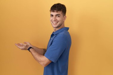 Young hispanic man standing over yellow background pointing aside with hands open palms showing copy space, presenting advertisement smiling excited happy 