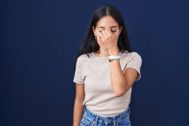 Mavi arka planda dikilen genç İspanyol kadın yorgun burnu ve gözleri ovuşturup yorgunluk ve baş ağrısı hissediyor. Stres ve hüsran kavramı. 