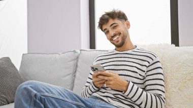 Neşeli genç bir Arap adam, koltuğunda rahat bir şekilde otururken akıllı telefonuyla sevinçle mesaj atıyor. Evinin sıcak, rahat oturma odasında mutluluk saçıyor.