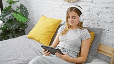 Yatak odasında dokunmatik ped kullanan, kulaklık takan genç sarışın kadın.