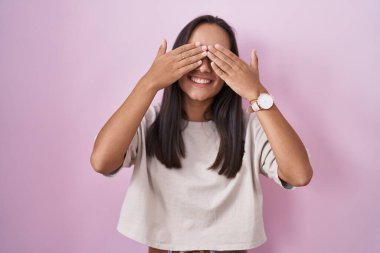 Young hispanic woman standing over pink background covering eyes with hands smiling cheerful and funny. blind concept. 
