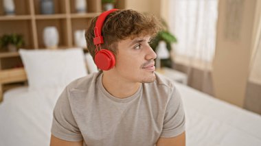 Genç, yakışıklı İspanyol adam rahat bir şekilde rahatlıyor, neşe içinde eğlenceli müzikler dinliyor, sade yatak odasında rahat bir yatakta oturuyor, mutluluğu ve güveni somutlaştırıyor ve sabahın tadını çıkarıyor.