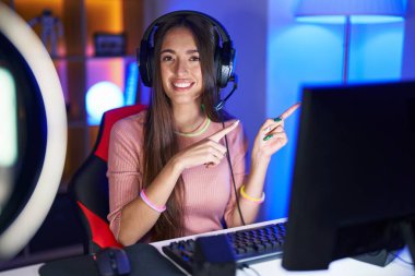 Genç İspanyol kadın video oyunları oynuyor gülümsüyor ve kameraya bakıyor iki eliyle ve parmaklarıyla kenarı işaret ediyor.. 