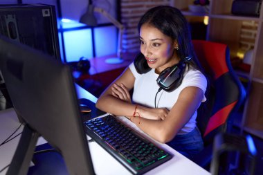 Young arab woman streamer smiling confident sitting with arms crossed gesture at gaming room