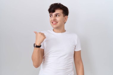 Gündelik beyaz t-shirt giyen, gülen yüzlü, gülümseyen ve baş parmağıyla yan tarafı gösteren genç, ikili olmayan bir adam.. 