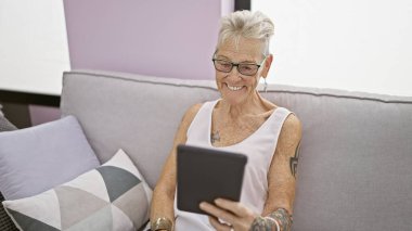 Kendine güveni tam, kısa gri saçlı, rahat koltukta dokunmatik ped kullanan olgun bir kadın. Orta yaşlı bir kadın oturma odasında teknolojinin tadını çıkarıyor..