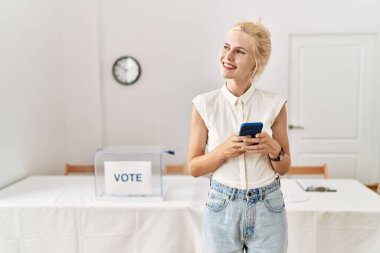 Seçmen okulunda gülümseyen akıllı telefon kullanan genç sarışın kadın.