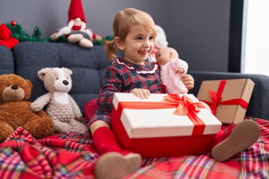 Sevimli beyaz kız elinde hediye ve bebek bebek evdeki Noel süslemesinde koltukta oturuyor.