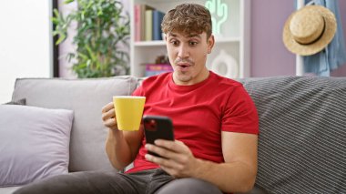 Şaşırmış genç İspanyol adam evde takılıyor, akıllı telefon kullanıyor, oturma odasındaki koltukta kahve içiyor.