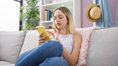 Evdeki kanepede oturan akıllı telefon kullanan genç sarışın kadın.