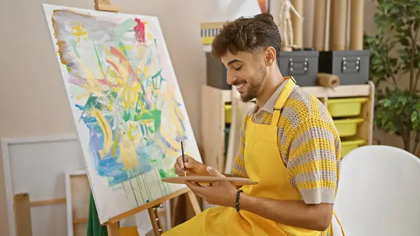 Confident young arab art student smiling as he expertly draws in a vibrant university art studio, harnessing his creativity with brush and palette