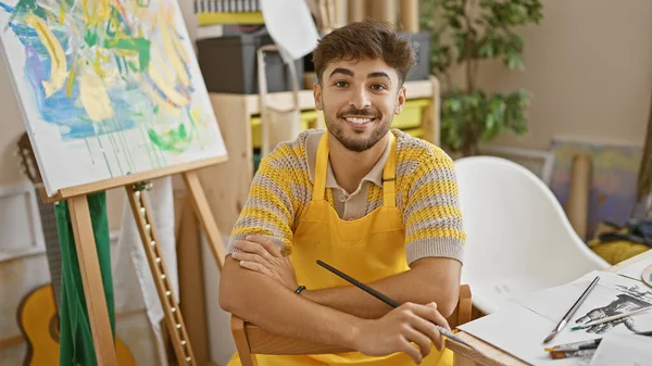 Confident, smiling young arab man artist sitting with arms crossed in his art studio, joyfully embracing his painting hobby
