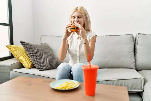 Young Blonde Woman Eating Hamburger Sitting Sofa Home — Stock Photo, Image