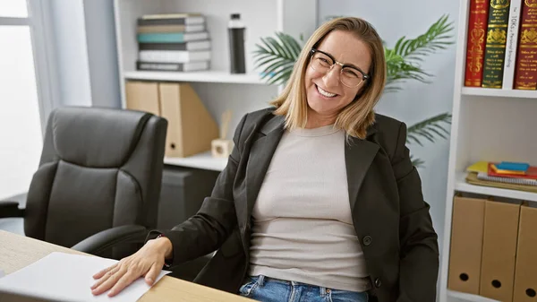 Joyful young woman boss enjoying successful business life, confidently smiling while sitting at office table. positive portrait of elegant blonde worker looking at camera, embodying happiness.