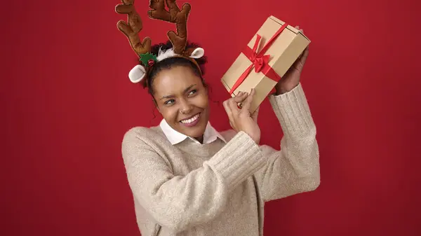Afroamerikanerin Hört Weihnachtsgeschenk Und Trägt Rentierohren Über Isoliertem Rotem Hintergrund — Stockfoto