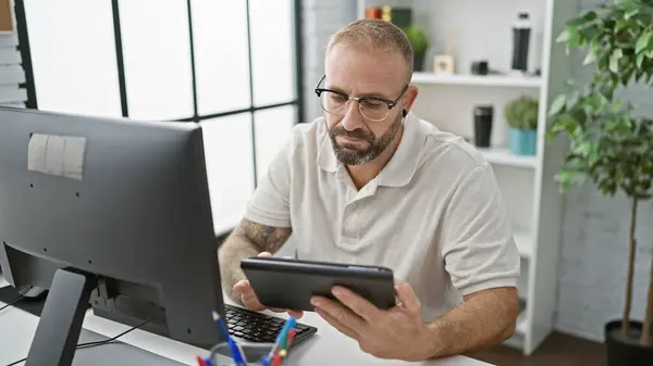 Handsome young man at work, relaxed yet serious business worker using touchpad and computer in office, embodying success in the indoor workplace.