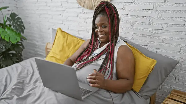 美丽而自信的非洲裔美国女人 早上在卧室里舒服地坐在舒适的床上 一边笑着 一边享受着笔记本电脑的乐趣 — 图库照片