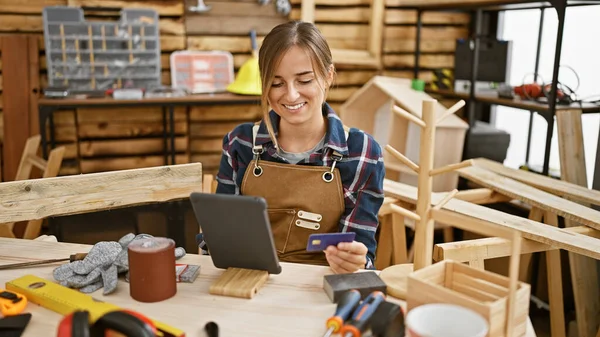 Attractive young blonde woman carpenter smiling while paying with credit card on touchpad amidst carpentry workshop