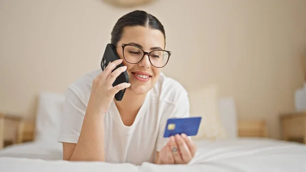 Young beautiful hispanic woman speaking on the phone shopping with credit card at bedroom