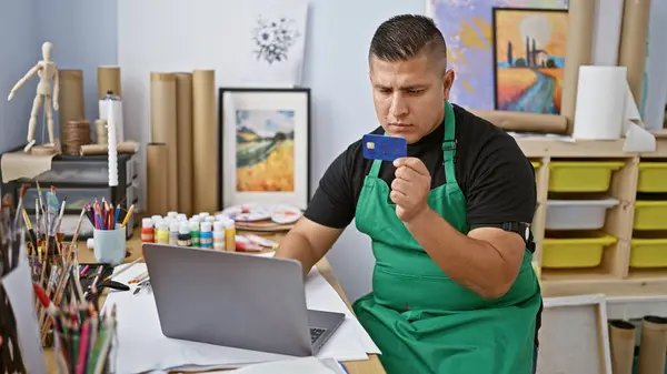 Hardworking latin artist, young man handling the responsibilities of his career with laptop and credit card in hand at art studio