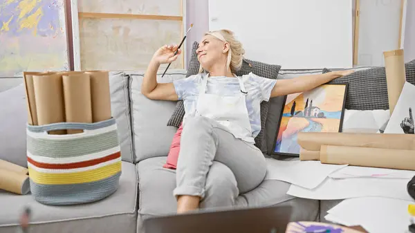 Joyful young blonde artist, paintbrush in hand, sitting on studio sofa, offers a heart-warming smile amidst her passion for painting