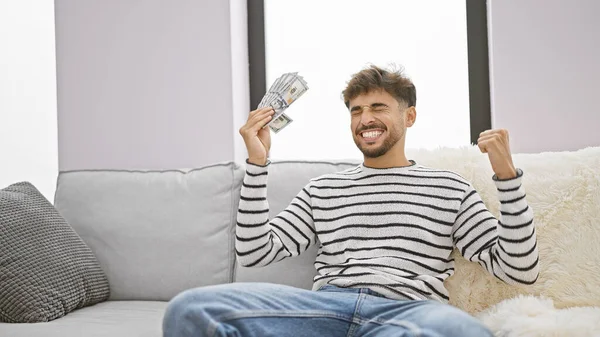 Joyful young arab man, cash in hand, basking in a winning moment on his cozy sofa, at home. his confident smile signals the positive expression of wealth - an investment win from us dollars