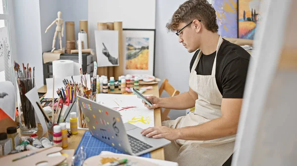 Handsome young hispanic man, an emerging artist, sitting at his art studio, enthusiastically learning drawing, using laptop and holding a smartphone.