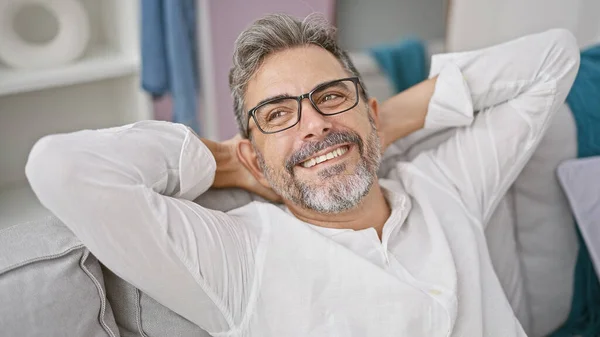 At home, a relaxed young hispanic man with grey hair is joyfully sitting on the sofa, hands on head conveying comfort and confidence