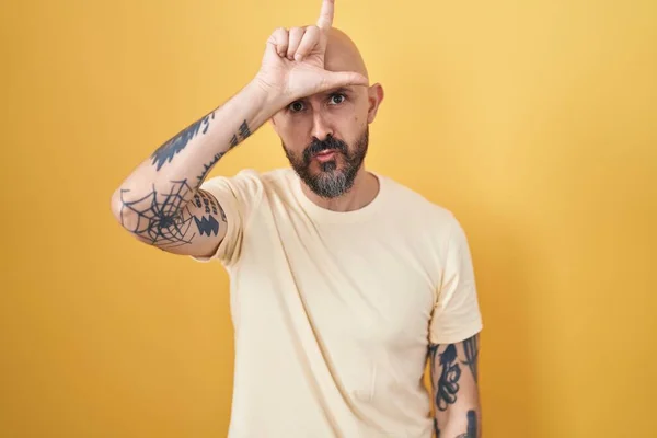 Hispanic man with tattoos standing over yellow background making fun of people with fingers on forehead doing loser gesture mocking and insulting.