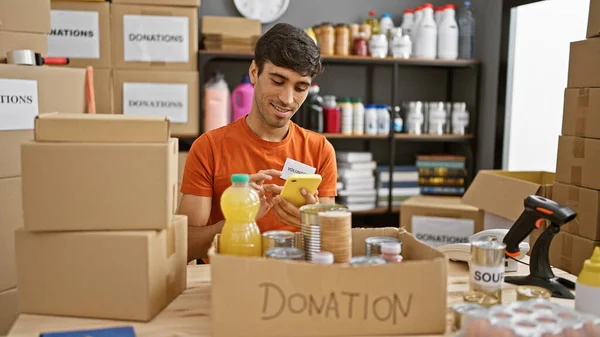Handsome young hispanic man volunteering at community charity center, smiling while texting donations updates on his smartphone, sitting amidst boxes of food donations in the warehouse