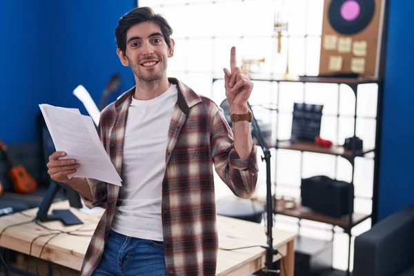 Young hispanic man reading music sheet at music studio surprised with an idea or question pointing finger with happy face, number one
