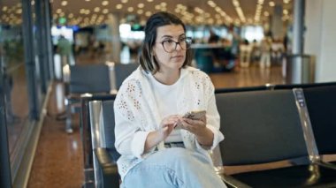 Güzel, genç, İspanyol bir kadın akıllı telefon kullanıyor. Ayakta duruyor ve havaalanında yürüyor.