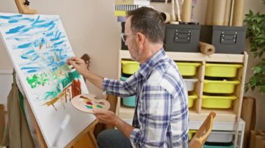 Gri saçlı, kendinden emin, pırıl pırıl gözleri olan, resim atölyesinde neşeyle resim yapan, yaratıcılık ve tutku saçan orta yaşlı bir adam.