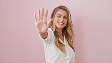Neşeli genç sarışın kadın, kendinden emin bir şekilde beş parmak gösteriyor! Hepsi giyinmiş, pembe bir arka planda tek başına durmuş, mutlu bir şekilde yukarıya bakıyor..