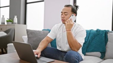 Genç Çinli adam dizüstü bilgisayar kullanıyor. Akıllı telefonla konuşuyor. Evdeki kanepede oturuyor.