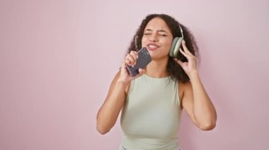 Havalı, kendinden emin, genç, güzel, kıvırcık saçlı, pembe izole arka planda neşeyle şarkı söyleyen ve dans eden, modern akıllı telefon cihazı ve kulaklıkla akılda kalıcı bir şarkı dinleyen İspanyol kadın.