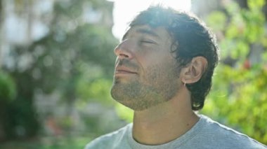 İspanyol genç adam gözleri kapalı, güneşli bir şehir parkında meditasyon yapıyor, özgürlüğün ve dengenin sembolü. Sarışın sakal, rahat bir portre, açık havada yaz güneşinin tadını çıkarmak..