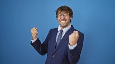 Heyecanlı takım elbiseli genç adam, mavi arka planda neşeli bir başarı çığlığıyla ünlü kazanan.