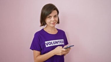 Kendine güveni tam, İspanyol bir kadın gönüllü, güzel gülümsüyor ve akıllı telefonundan hayırsever bağışları mesaj atıyor. Pembe bir duvar arkaplanı üzerinde izole.
