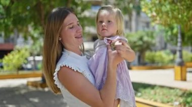 Kendine güveni tam bir anne, kollarını kızına dolamış, neşeyle gülüyor, güneşli parkta duruyor, pozitif enerji ve mutluluk saçıyor.