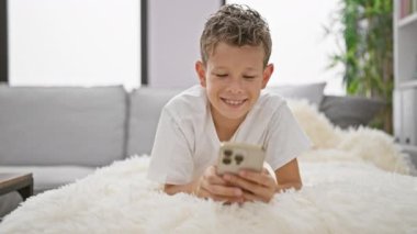 Sevimli sarışın çocuk, neşe dolu, akıllı telefonunu neşeyle kullanıyor, evde, kanepede yatarken mesajlaşıyor.