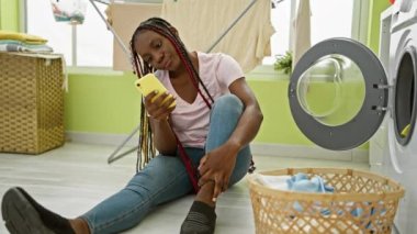 Güzel Afrikalı Amerikalı kadın çamaşırhanede oturmuş, ev işleri arasında akıllı telefonuyla mesajlaşırken gülümsüyor.
