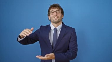 Takım elbiseli neşeli genç adam kocaman mutlu bir gülümseme takınıyor ve elleriyle kocaman bir işaret veriyor. Serin, mavi, izole bir zemin üzerinde - 