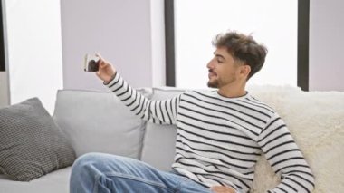 Kaygısız, genç Arap adam evde, mutlu bir şekilde anın tadını çıkarıyor, mutluluğunu ifade ediyor, oturma odasındaki kanepede otururken akıllı telefonuyla selfie çekiyor.