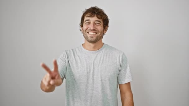 カジュアルなTシャツの陽気な若者は 自信を持って2本の指で勝利のサインを見せ 白い壁の背景に隔離されたカメラを見ながら勝利の笑顔を点滅させます — ストック動画