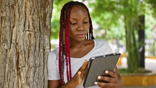 自信的非洲裔美国女人 美丽而快乐 靠在公园的树上 沐浴在阳光下 在户外使用她的触摸板时 闪烁着清凉而快乐的微笑 — 图库视频影像