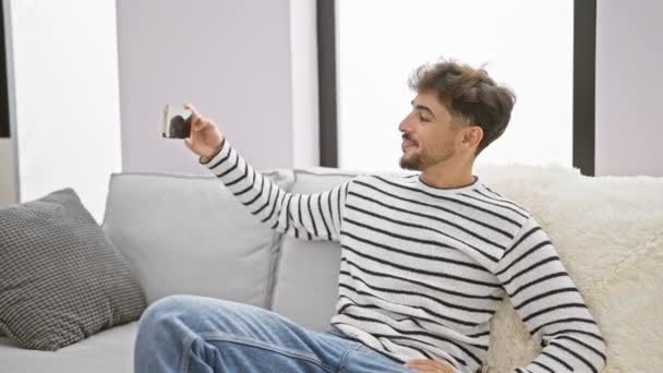 无忧无虑 年轻的阿拉伯男子在家里 快乐地享受着这一刻 表达着喜悦 一边坐在客厅的沙发上一边用智能手机自私自利 — 图库视频影像
