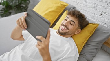 Yatak odasının rahatlığında, genç, gülümseyen bir Arap adam yatağa uzanıyor, dokunmatik bilgisayarında bir video seyrediyor, her anın tadını çıkarıyor..