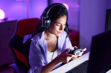 Young beautiful hispanic woman streamer playing video game using joystick at gaming room